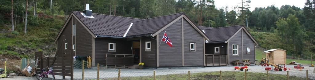 Brun bygning og litt av uteplassen til Bergsøy barnehage. Norsk balkongflagg på et hjørne ved inngangspartiet. - Klikk for stort bilde