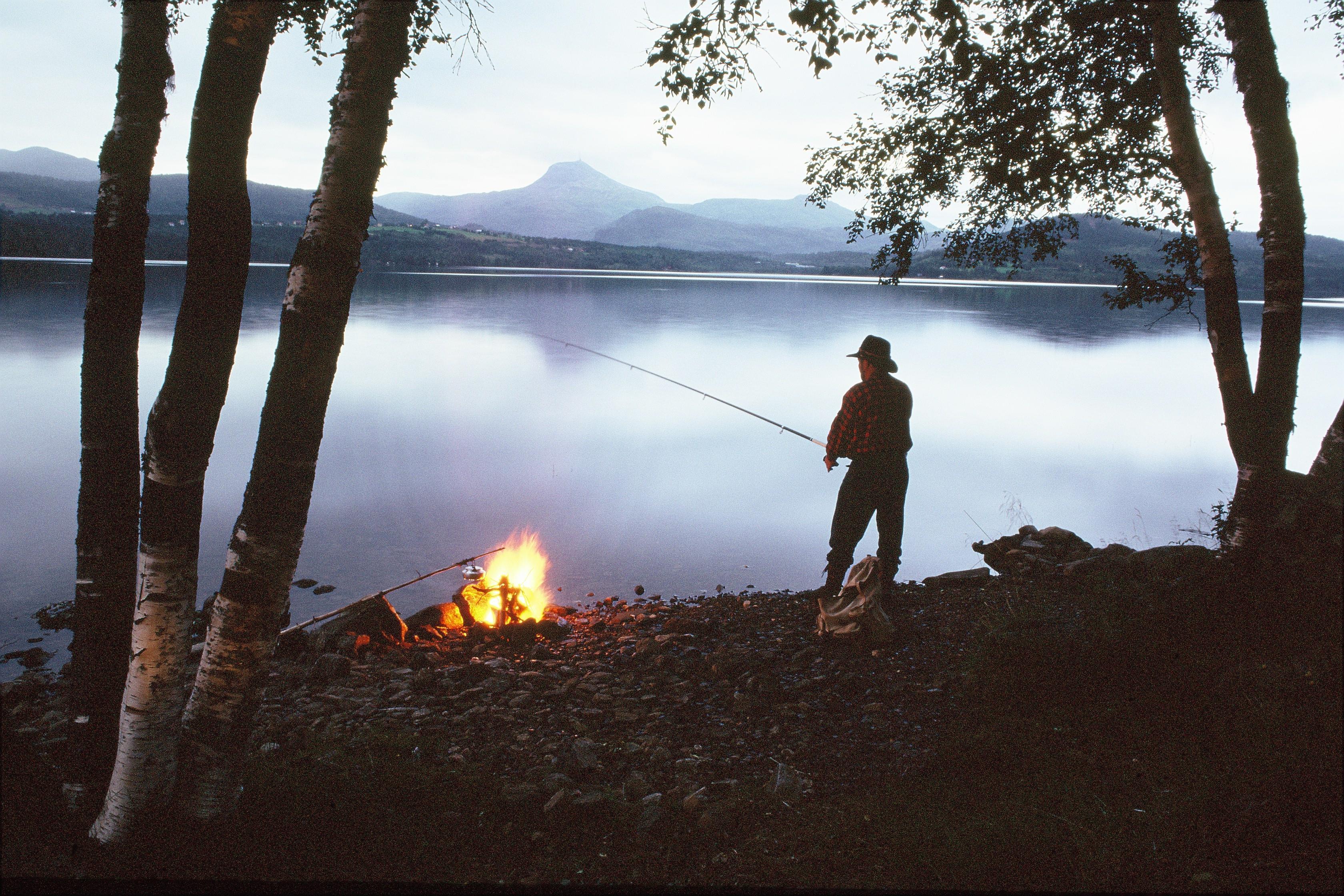 kveldstemnig med stille vatn, Mann med hatt står og fisker, bål i vannkanten med kaffekjele som henger over bålet - Klikk for stort bilde